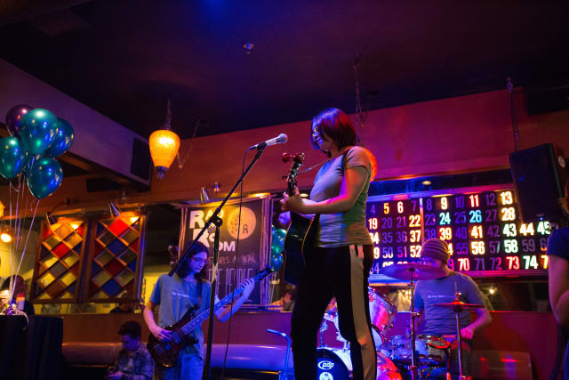 Youth musicians performing on stage at the Love Bomb 2017 event, with a guitarist singing into a microphone, a bassist, and a drummer, surrounded by colorful balloons and atmospheric lighting.